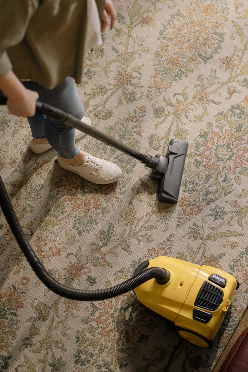 Person cleaning a floral carpet with a yellow vacuum cleaner indoors.